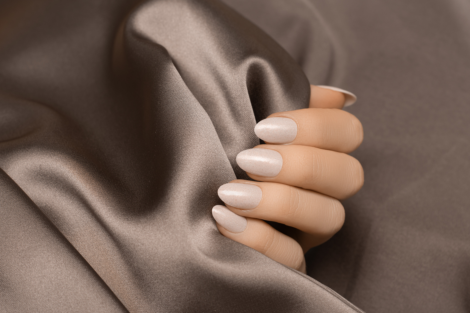 Female hand with white nail design. White glitter nail polish manicure on brown fabric background